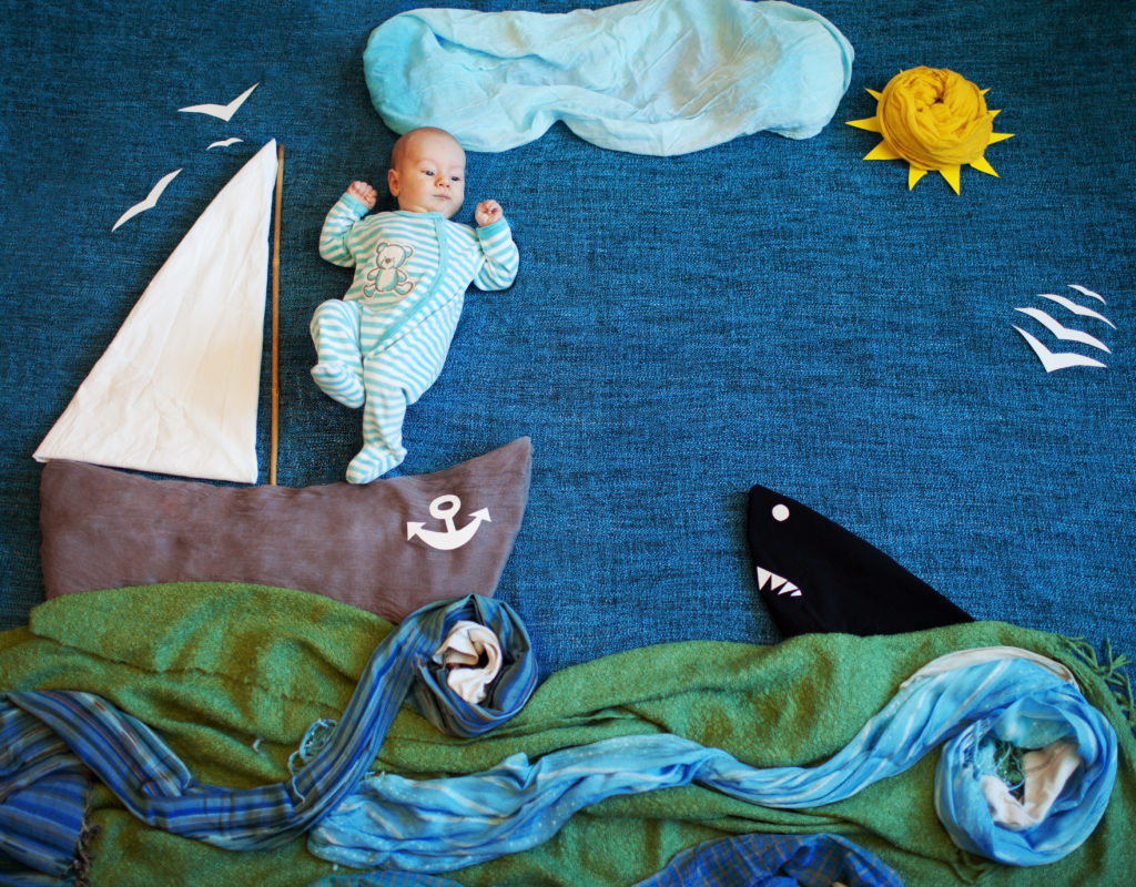 baby girl laying on a blanket that looks like the blue sky. There is a material cloud above her, a yellow sun, and a material boat with waves. The entire background is fabric shaped to look like a sea.