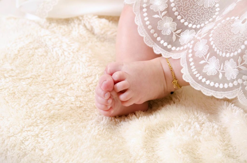 close up of a newborn feet, a gold religious charm clasped around the ankle. Baby is wearing baptismal outfit.